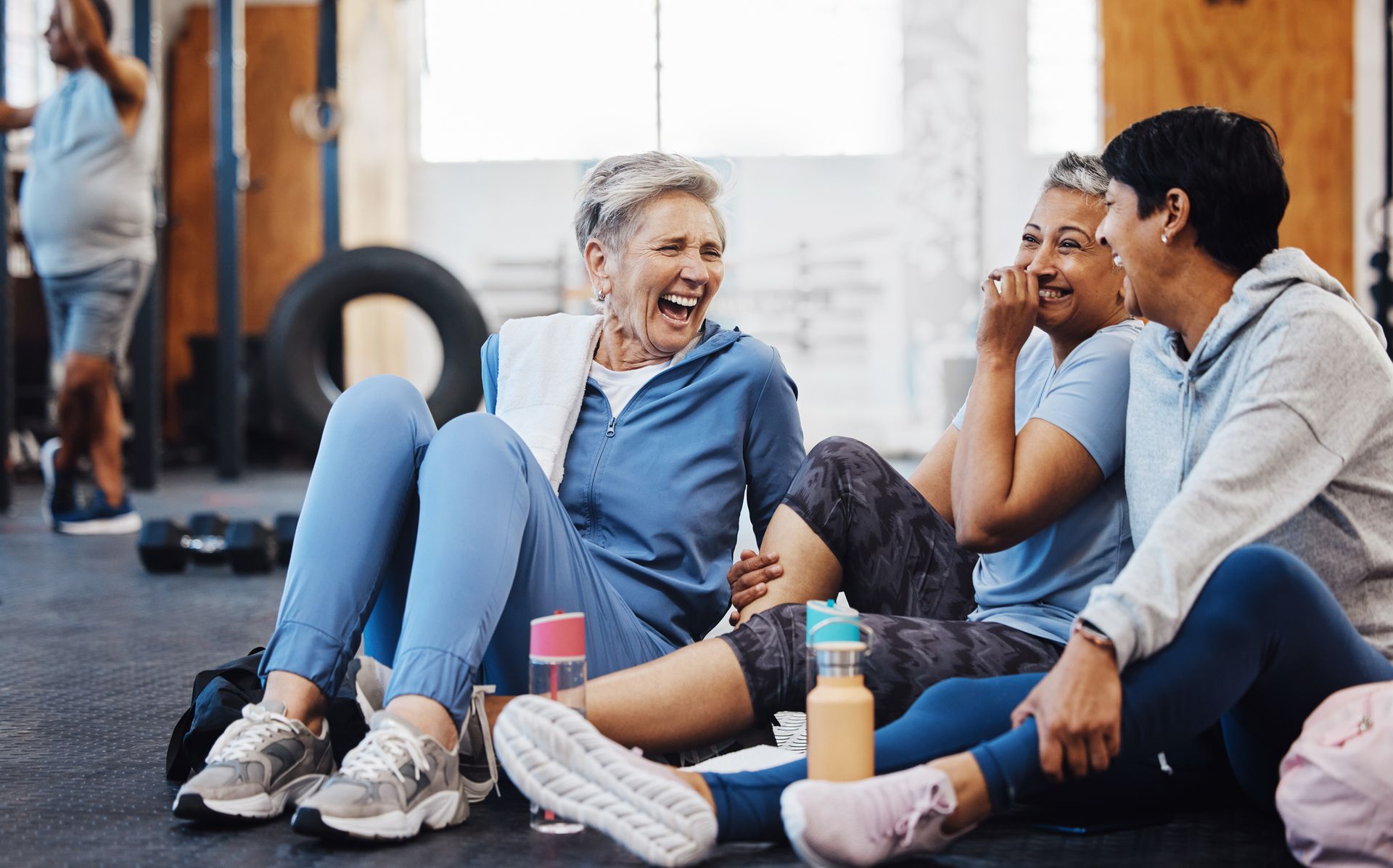 Woman sitting on bed, holding a glass of water, stretching with a smile in a bright bedroom. Group of people in workout clothes laughing together on a gym floor.