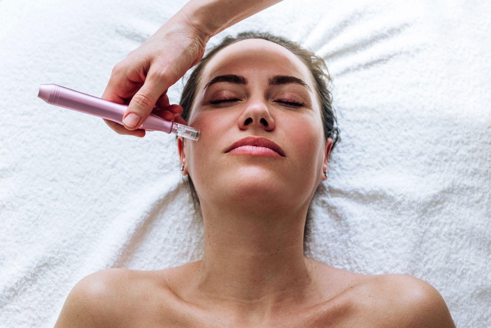 Woman receiving facial treatment, pink device held to her cheek on a white towel. Woman receiving facial treatment, pink device held to her cheek on a white towel.