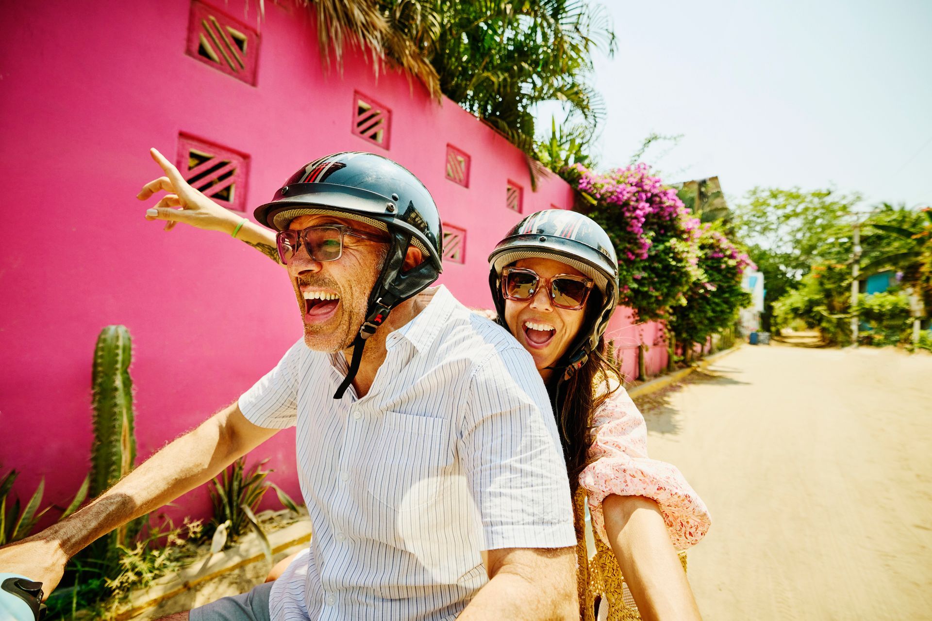 Chalkboard reads Couple on a scooter in front of a pink wall, both wearing helmets, smiling, and pointing.