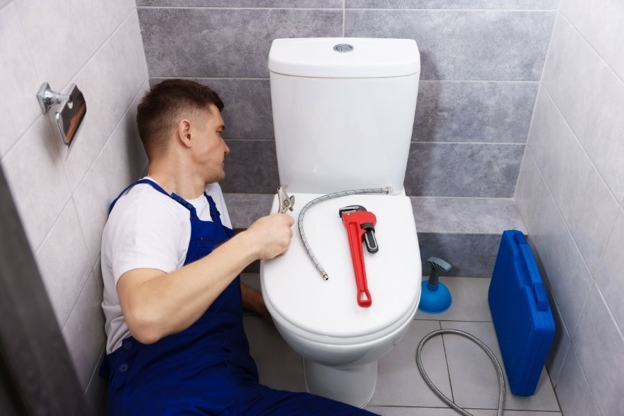 A homeowner kneeling next to a toilet checking the floor and supply line for moisture and corrosion