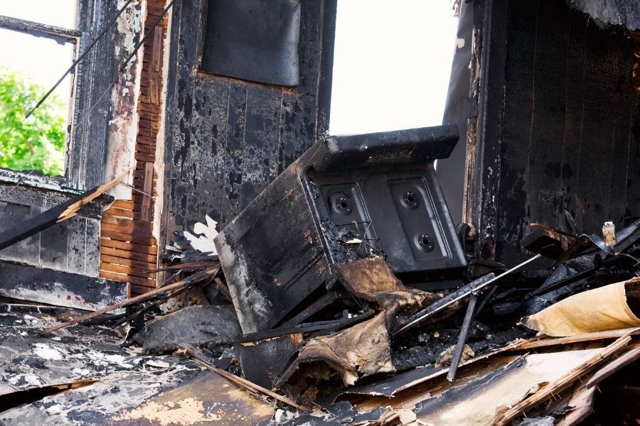 Burned household item among charred debris and ash near a window inside a fire-damaged home