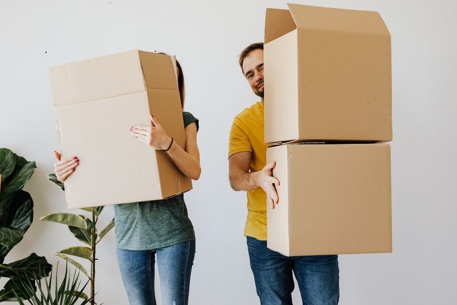 A couple holding cardboard moving boxes inside a home with a leafy plant visible in the corner