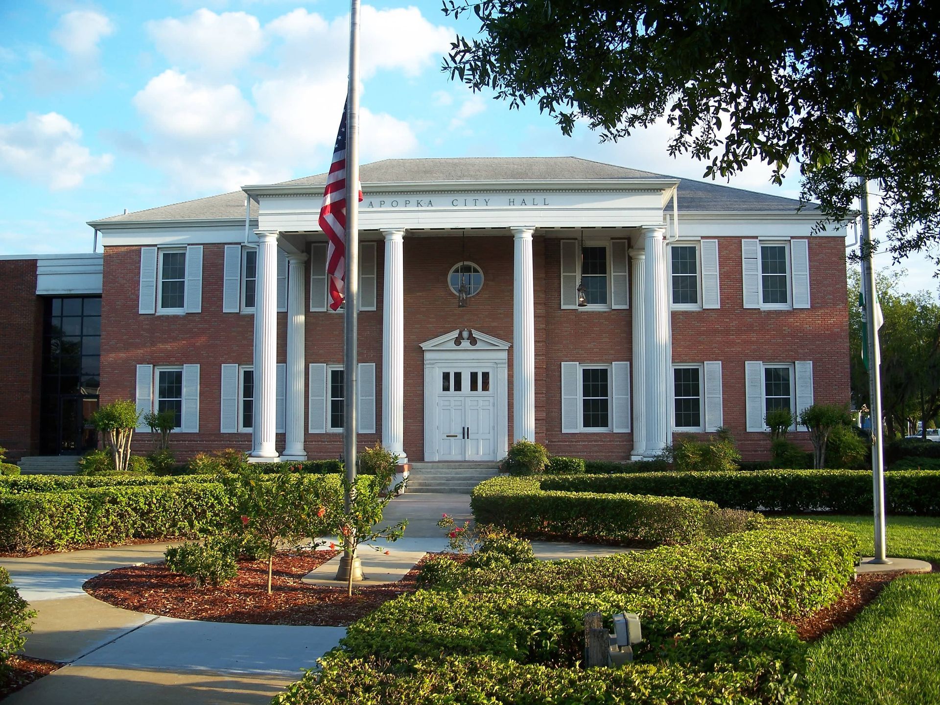 A brick-red building with white accents and surrounded by greenery in Apopka, Florida, features an American flag on a nearby flagpole.