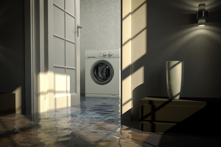 Water pooling on a laundry room tile floor around the base of a front-loading washing machine, with visible moisture line on adjacent drywall]