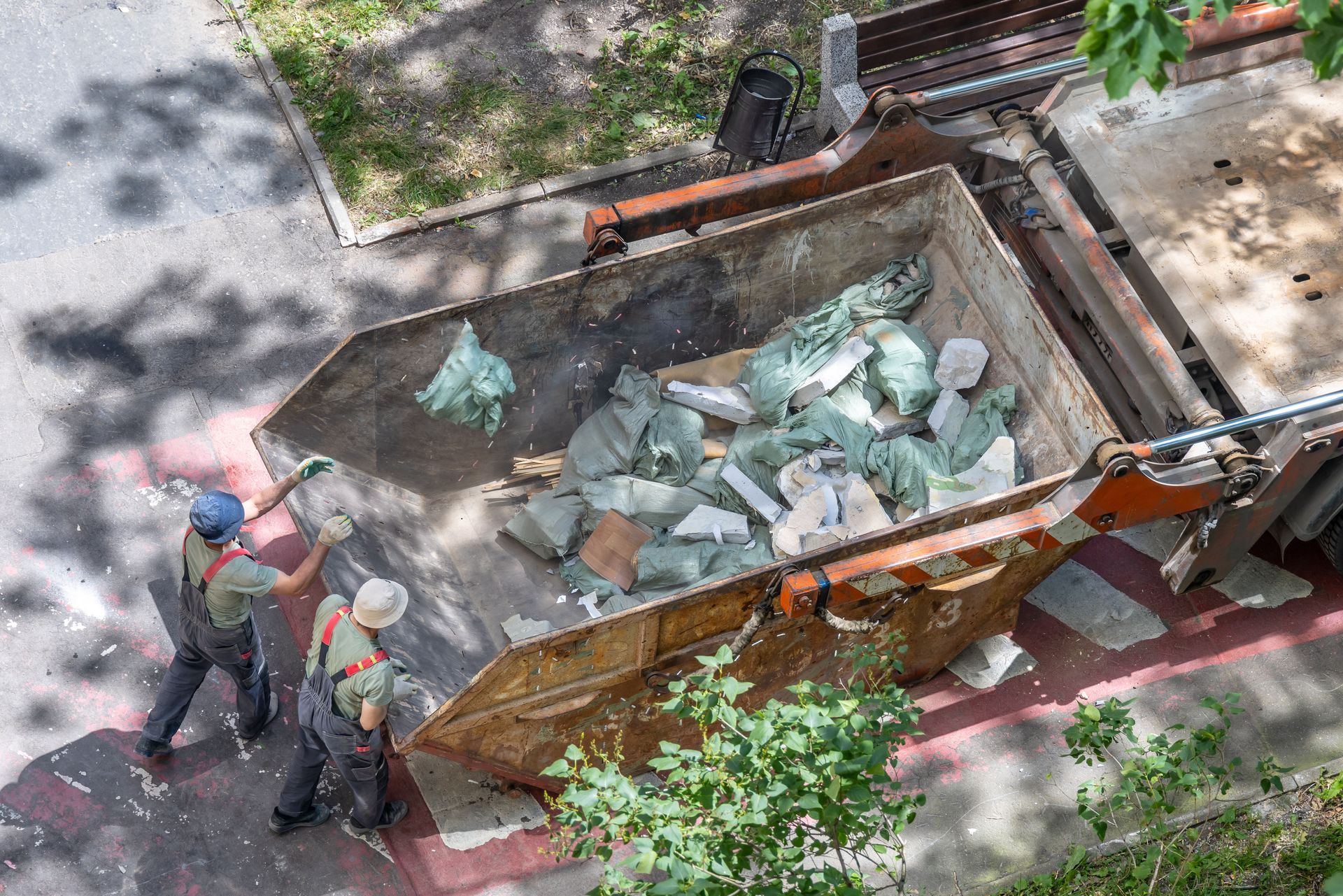 Workers emptying debris into a large dumpster on a sidewalk beside trees