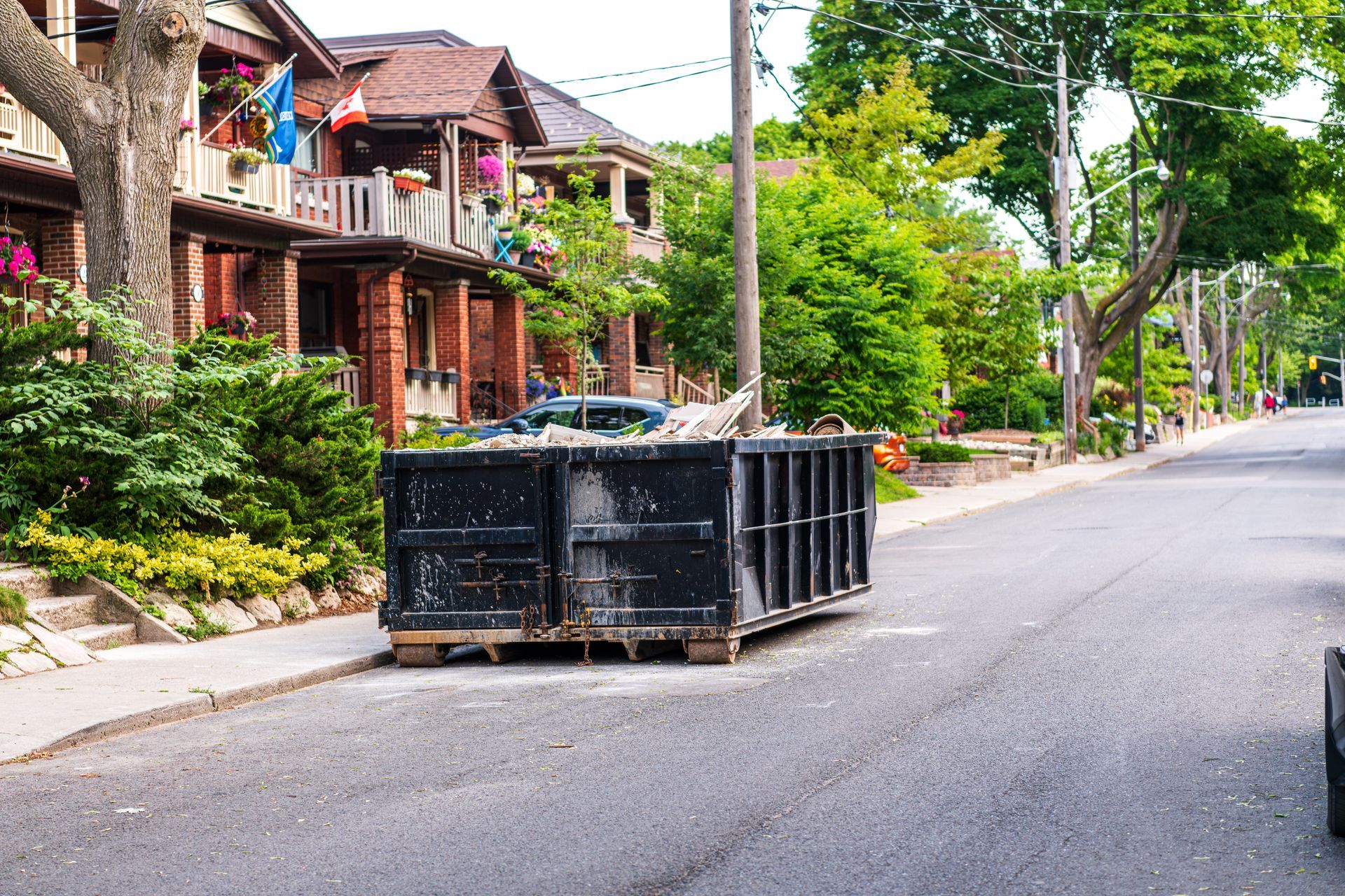 Two black dumpsters on a quiet residential street lined with brick houses and trees