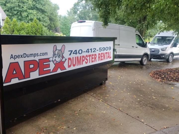 Large black dumpster on a residential street in front of brick houses and trees