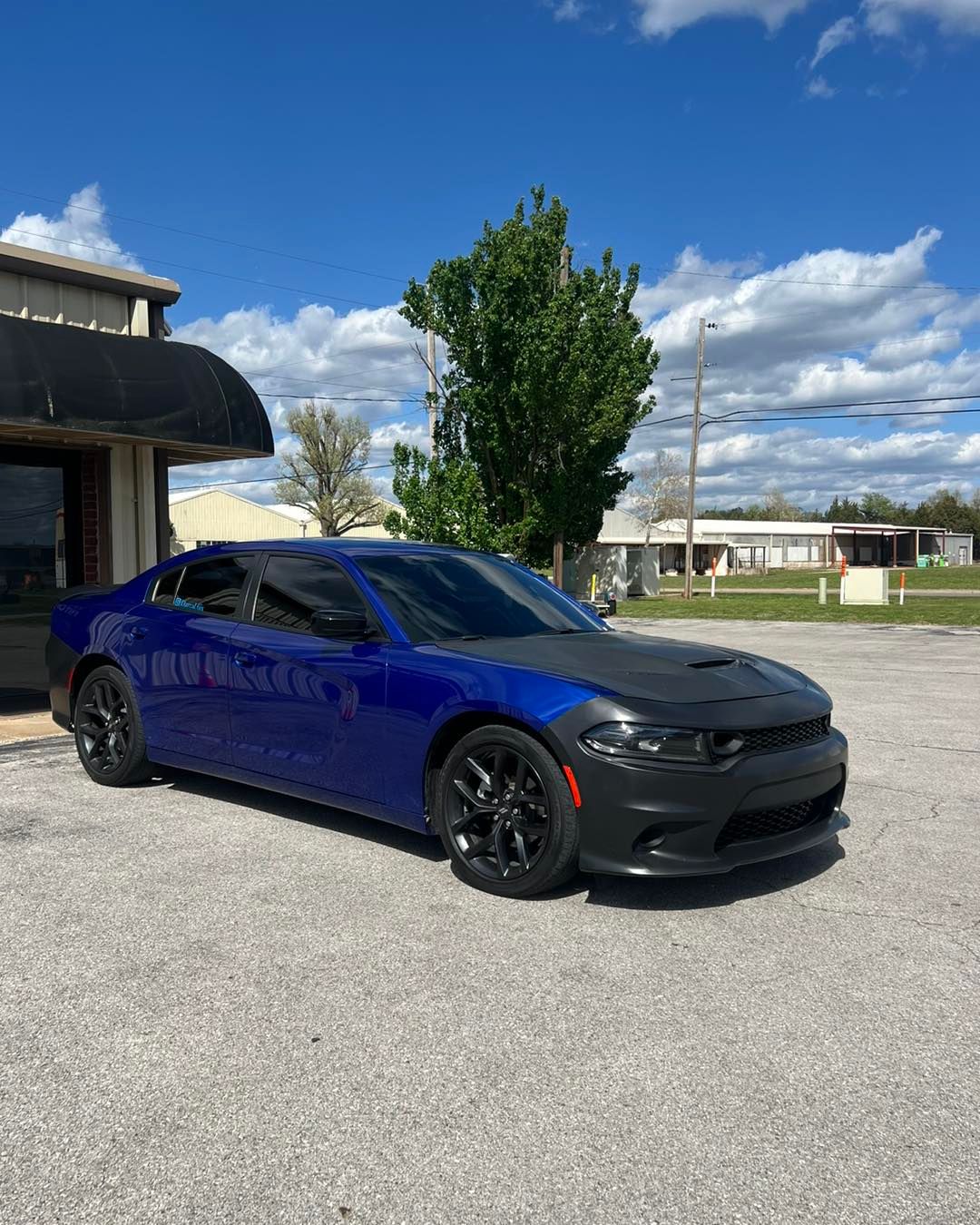 Blue Dodge Charger with black hood cover and black wheels parked in a lot under a blue sky.