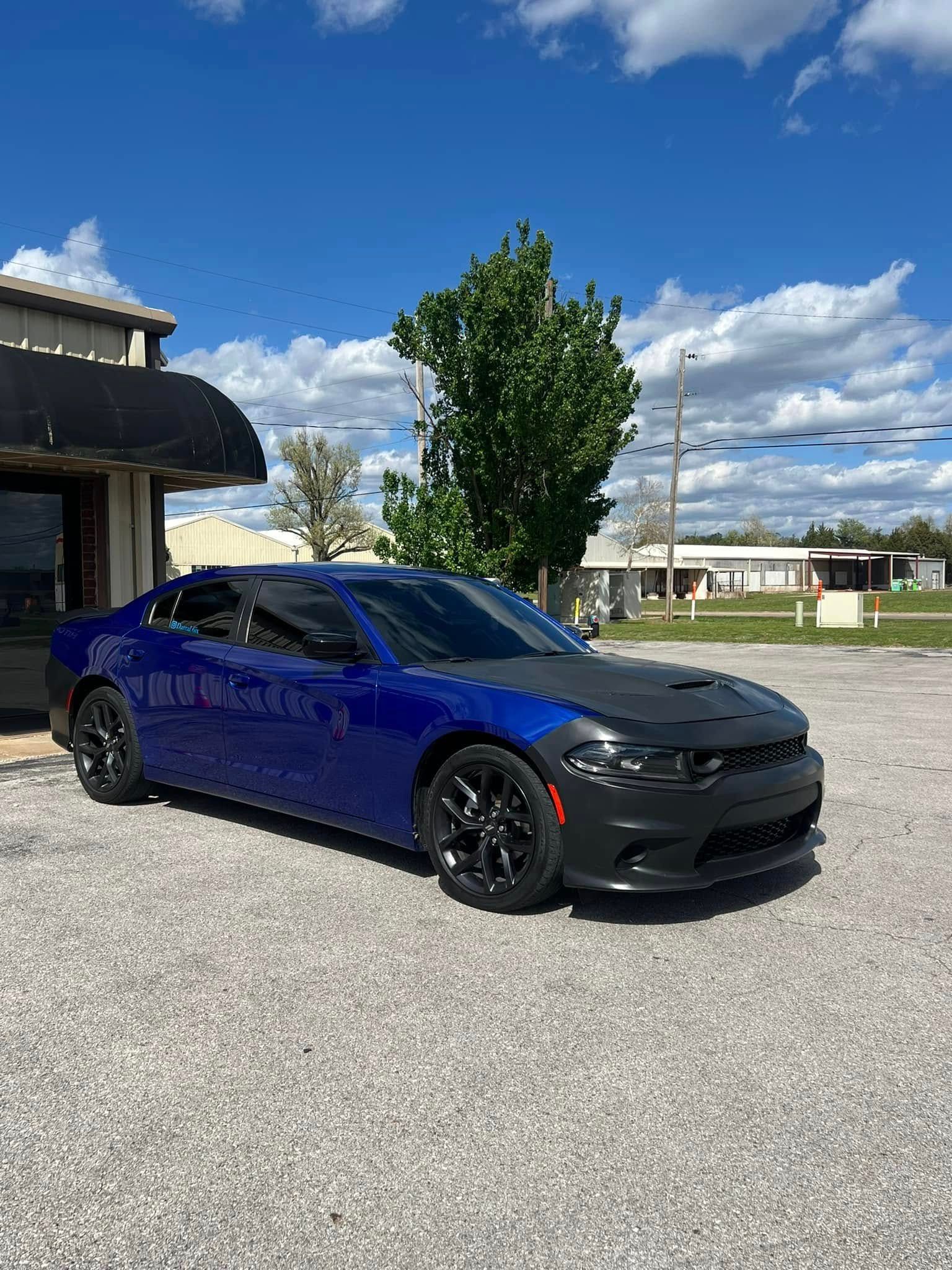 Blue Dodge Charger parked in front of a building. Black hood cover, black wheels, and bright sky.