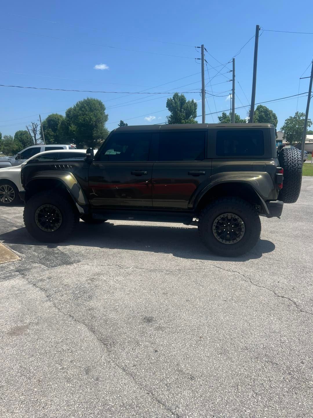Green Ford Bronco SUV parked on asphalt. Black wheels, spare tire, tinted windows, under blue sky.
