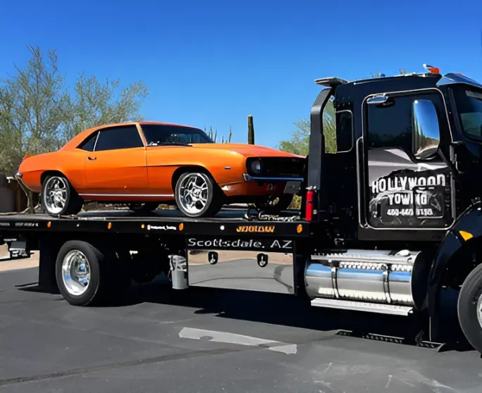Orange classic car on a black tow truck in a parking lot under a clear blue sky