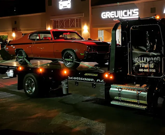 Orange classic car on a tow truck at night outside Greulich's garage.