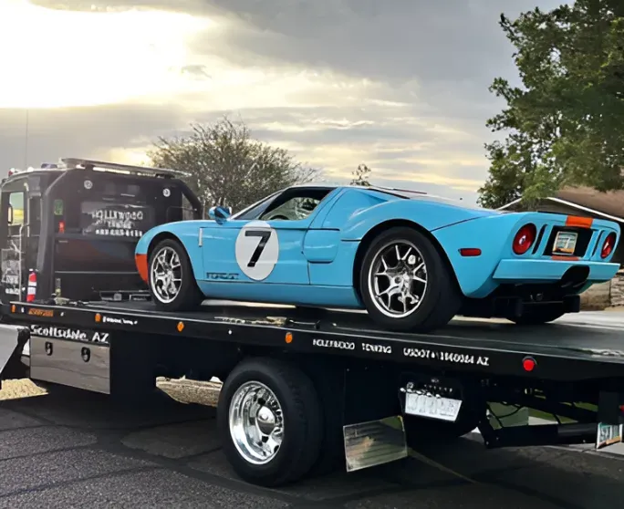 Blue race car on a black trailer, parked outdoors at sunset with another vehicle nearby.