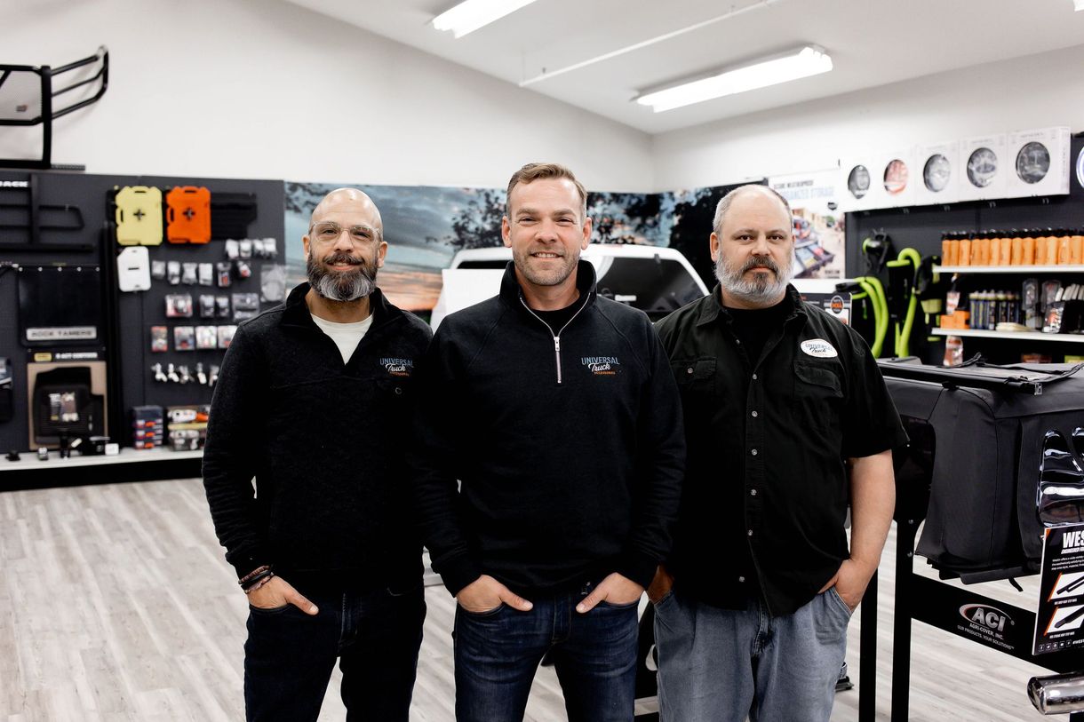 Three men pose in a store with vehicle accessories. Two are wearing black tops, and the third is in a dark shirt and jeans.