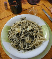 Pasta al pesto e aglio su un piatto bianco, con bicchiere di vino, bottiglia e coltello sullo sfondo.