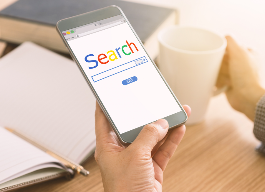 Hand holding a smartphone showing a search page beside an open notebook and coffee cup