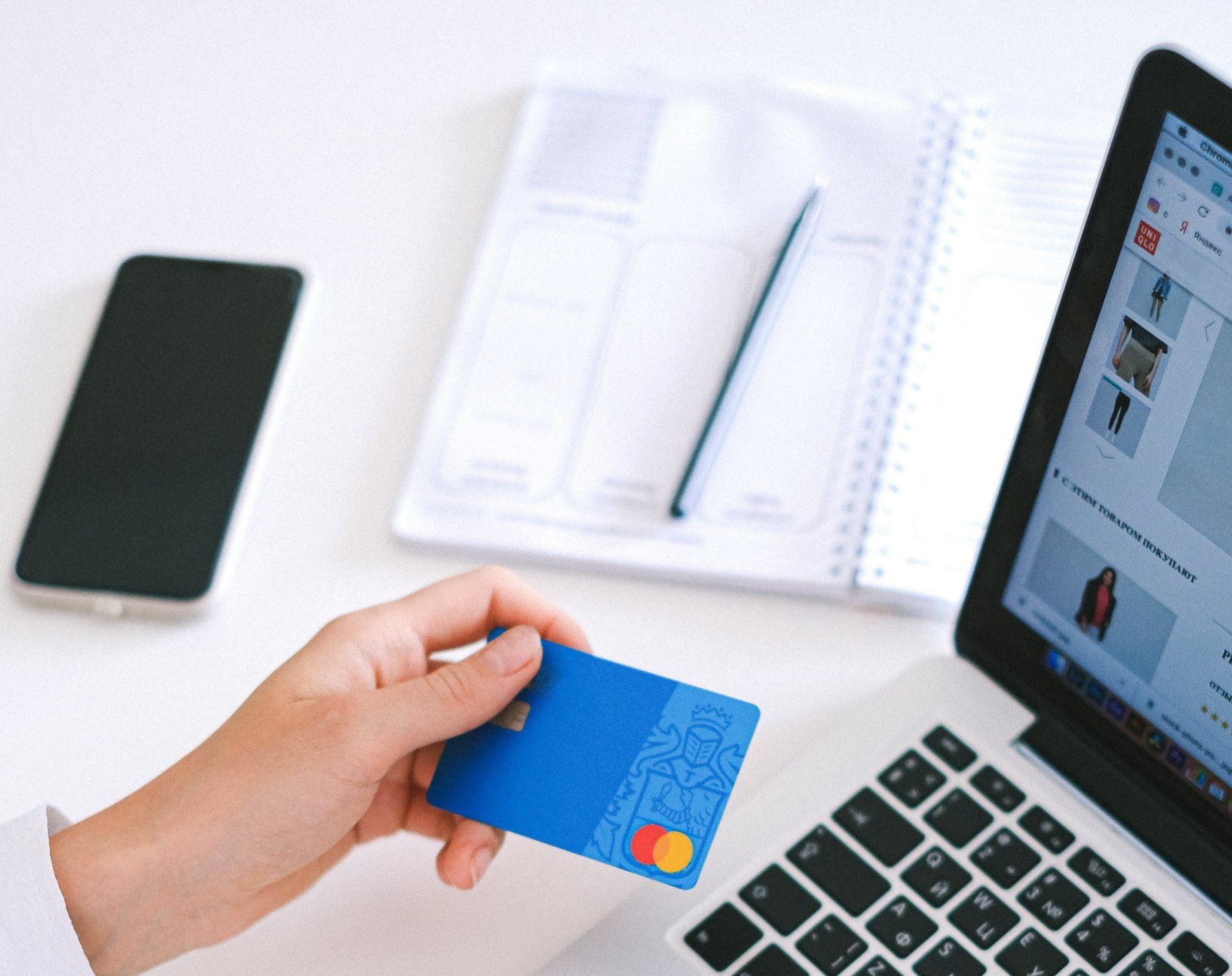 Hand holding a blue credit card near a laptop keyboard, with phone and notebook on a desk