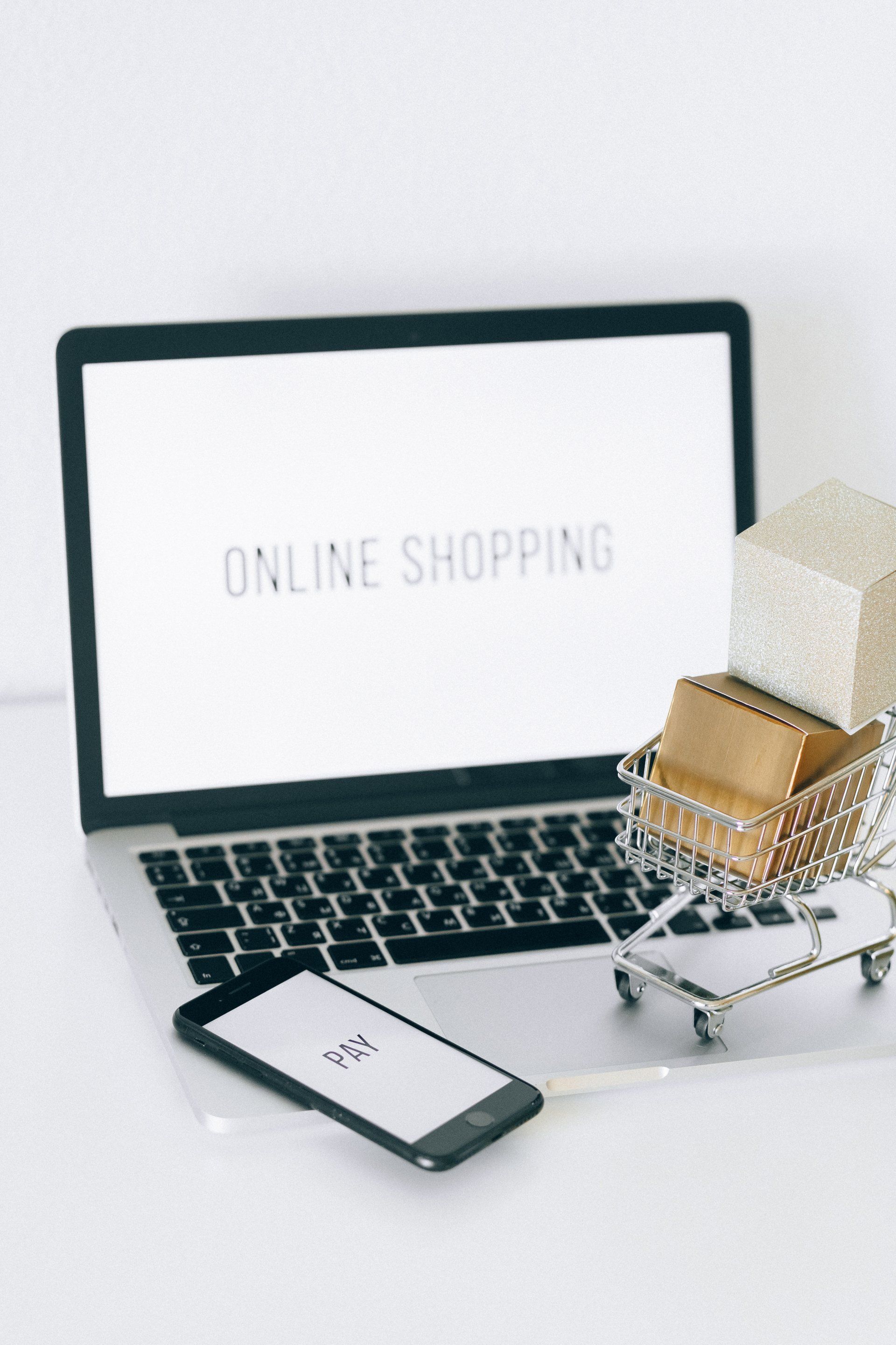 Laptop and smartphone displaying online shopping, with a gift box and shopping cart on a white desk.