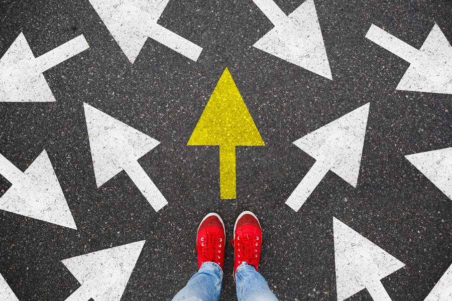 Feet in red sneakers facing a yellow forward arrow among white arrows on asphalt