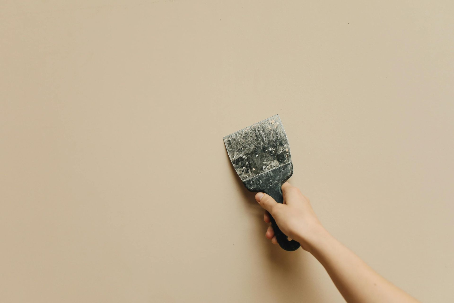 Hand holding a paint scraper against a beige wall