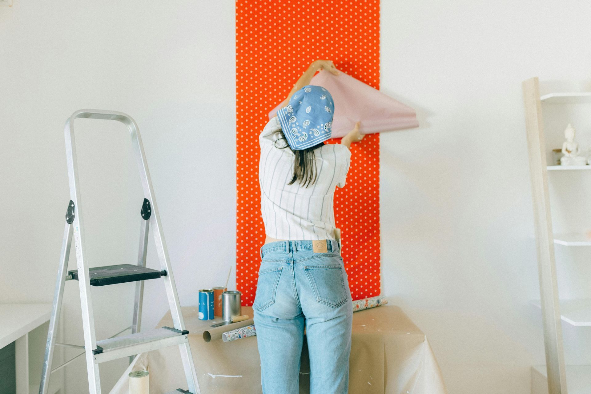 Person in a room wallpapering an orange accent wall beside a step ladder and paint cans