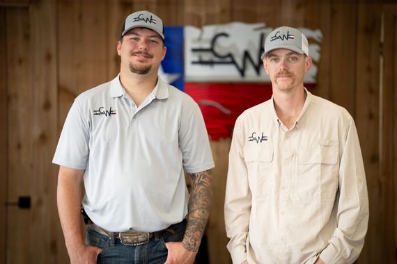 Two men in work shirts and caps standing in front of a wood wall and red logo sign.