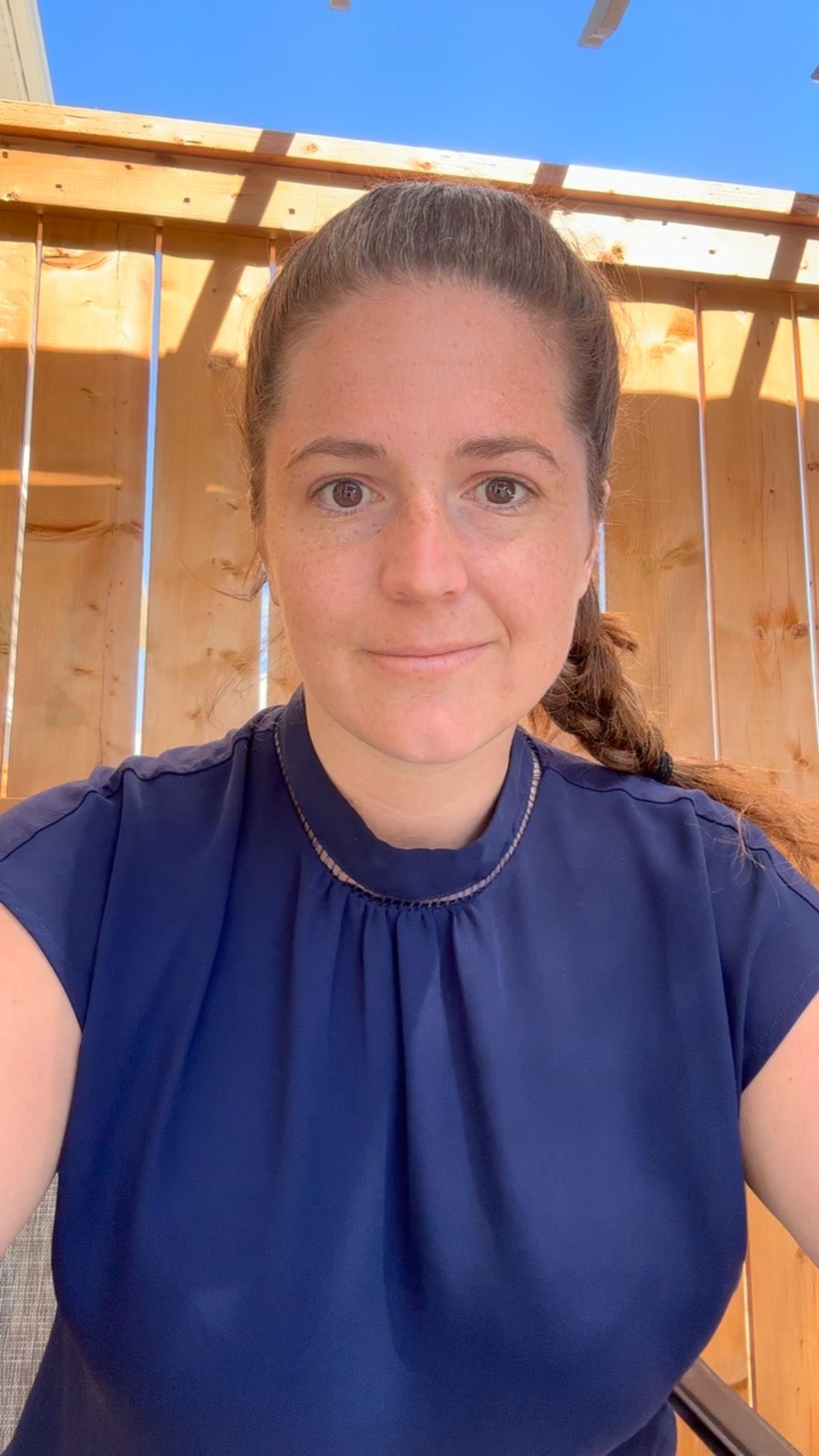 Woman with brown hair in a blue blouse smiles, outside with wooden beams and a blue sky.