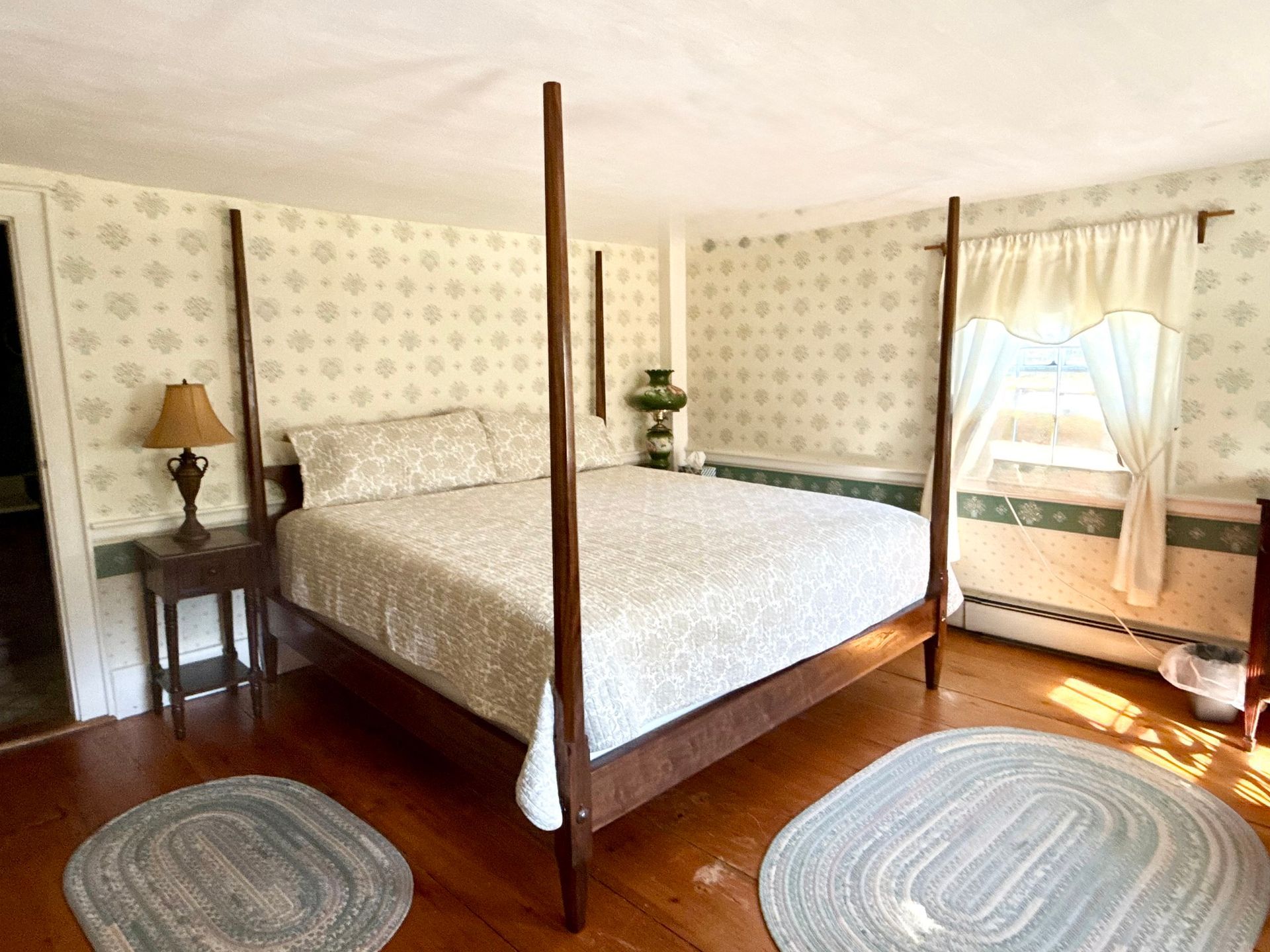 Four-poster bed in a sunlit bedroom with floral wallpaper, wooden floors, rugs, and a window seat