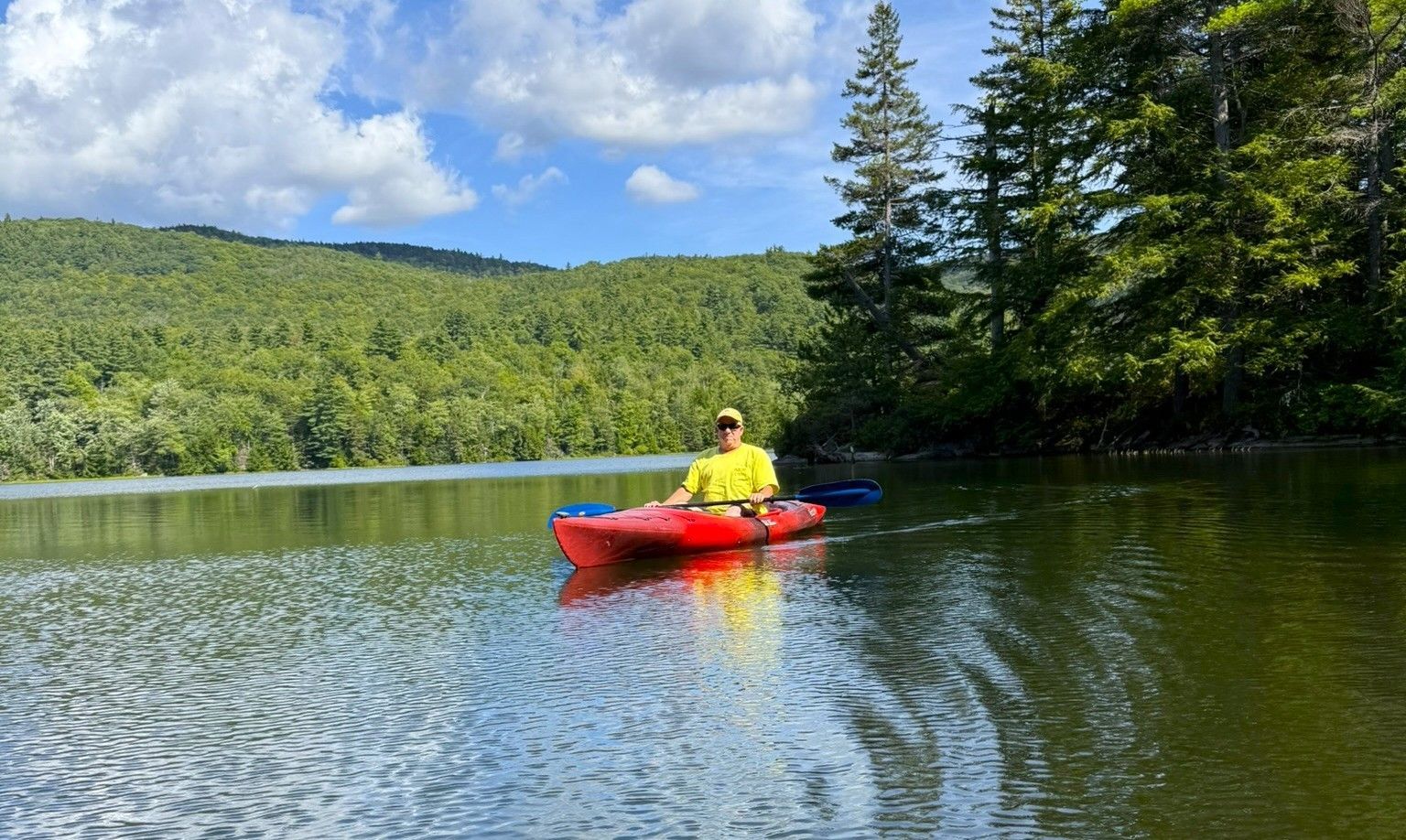 Person in a red kayak on a calm lake, with green forested hills and cloudy sky in the background