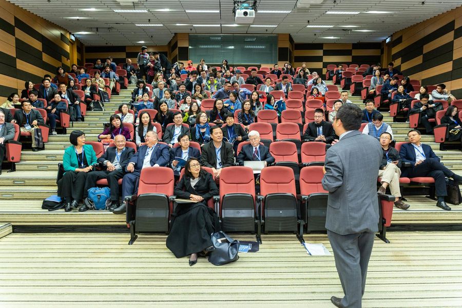 A speaker stands before a large, tiered lecture hall filled with an audience seated in rows of red chairs.