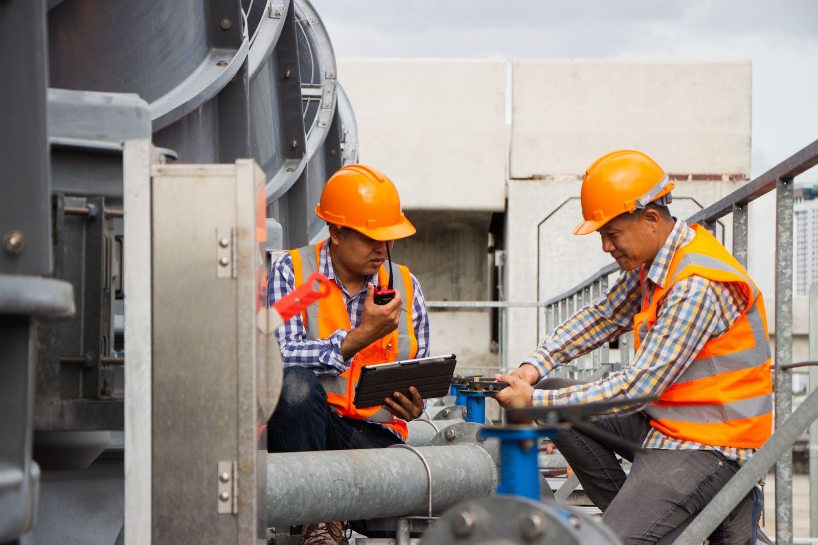 Two construction workers are working on a pipe and looking at a tablet.
