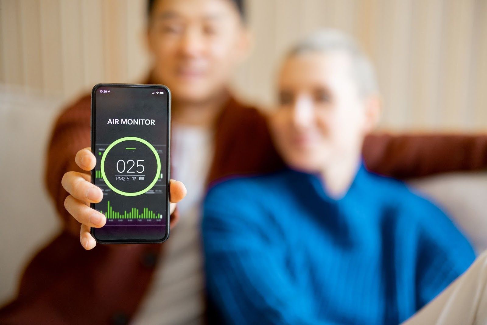 A man is holding a cell phone in front of a woman sitting on a couch.