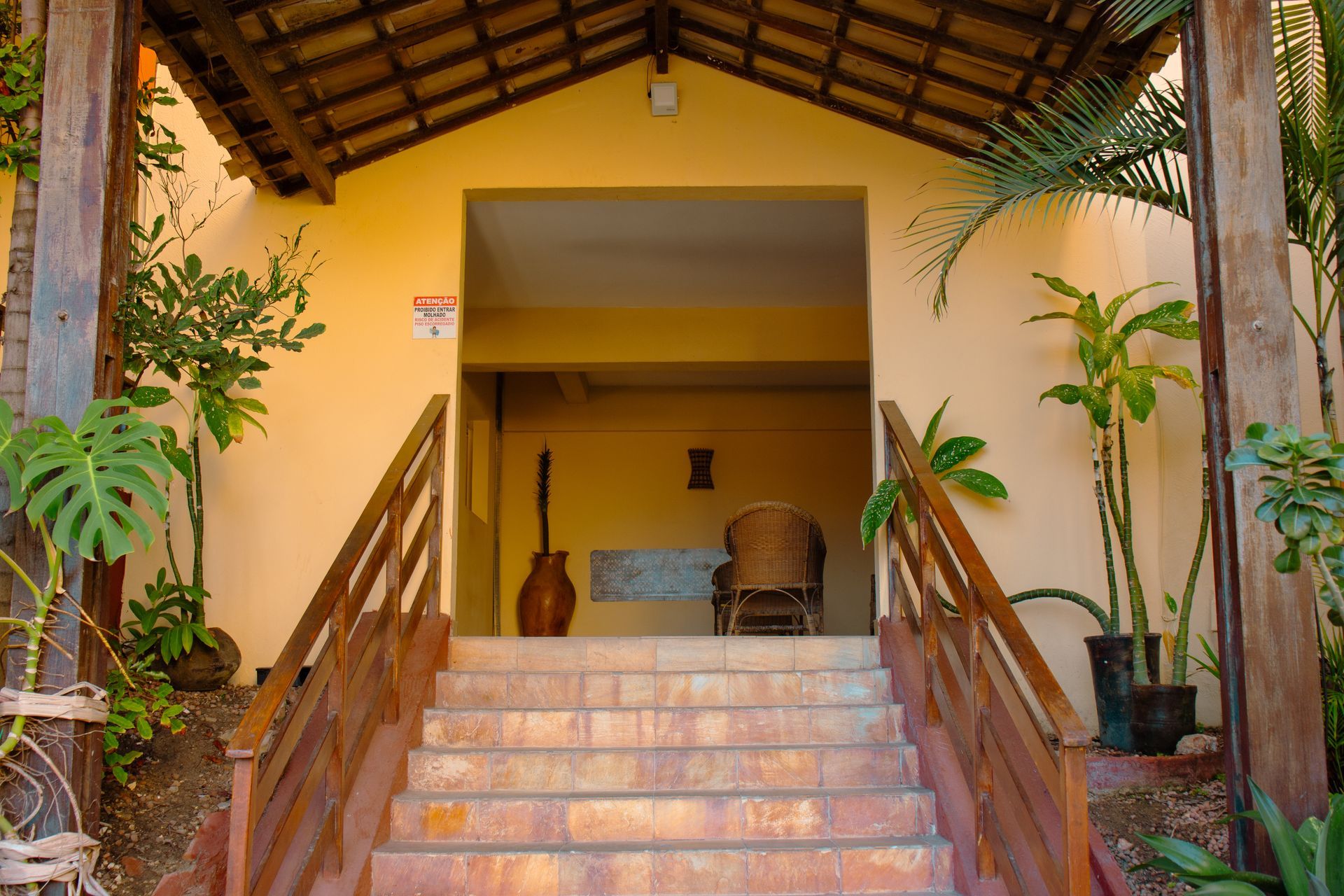 Entrada coberta com escadas, vasos de plantas e corrimãos de madeira que levam a um prédio amarelo.