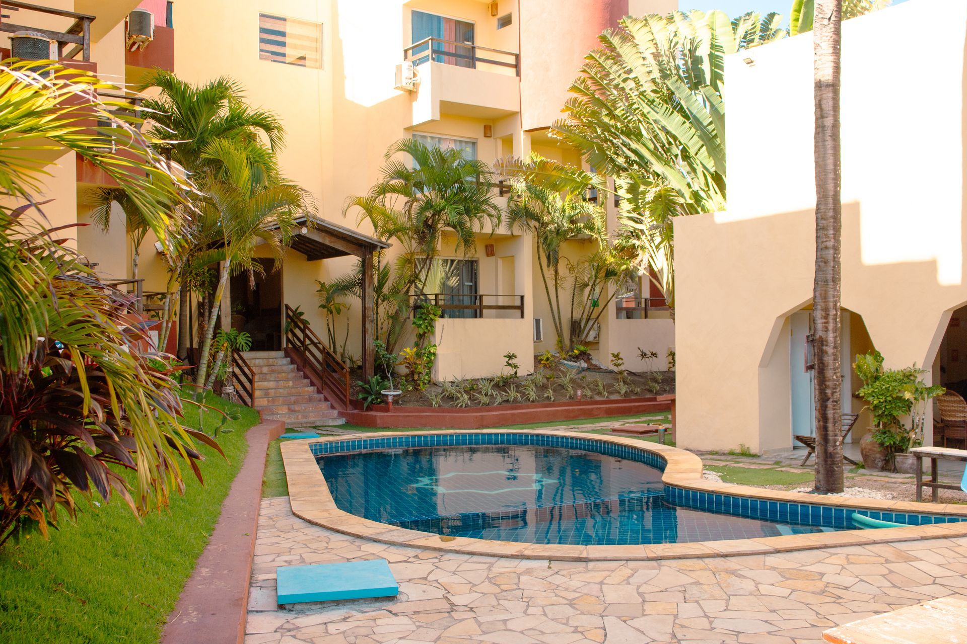 Piscina no pátio com água azul, palmeiras e prédios de apartamentos bege sob um sol quente.