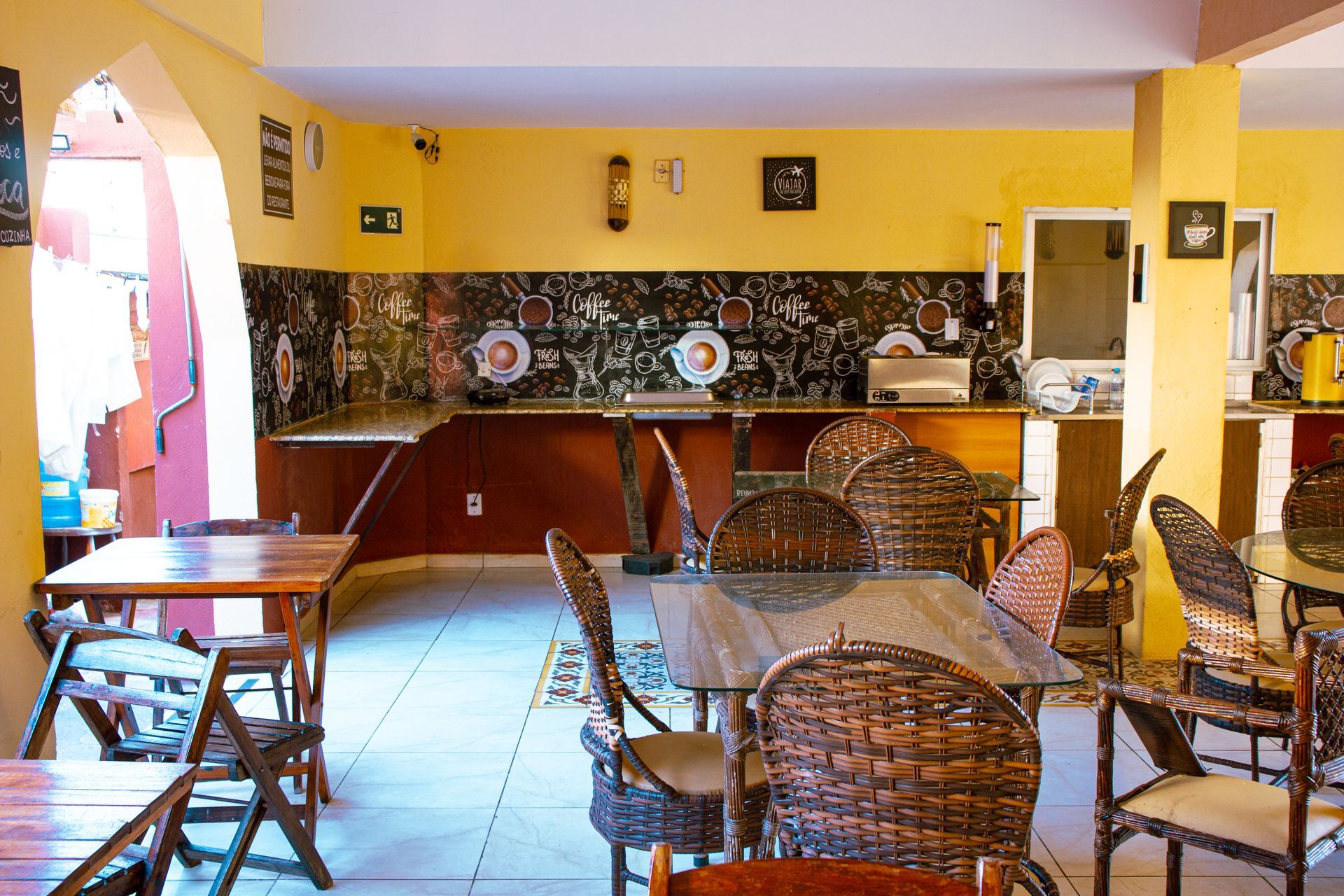 Café vazio com cadeiras de vime, mesas de madeira e um balcão amarelo com pratos decorativos.