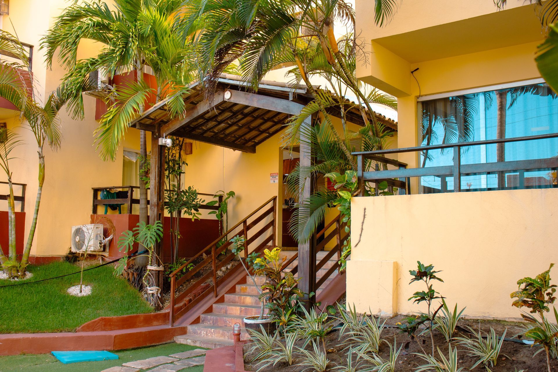 Entrada para o pátio com escadas, plantas tropicais e um prédio amarelo com janelas de vidro azulado.