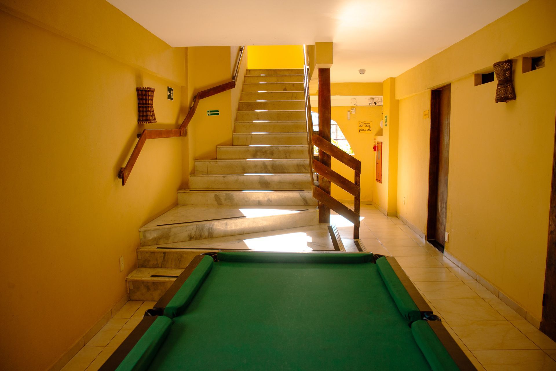 Corredor amarelo com escadas e uma mesa verde em primeiro plano, iluminado pela luz do sol.