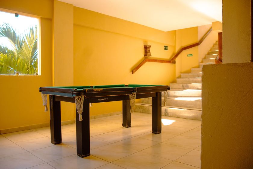Mesa de bilhar em uma sala interna ensolarada com paredes amarelas e uma escada.