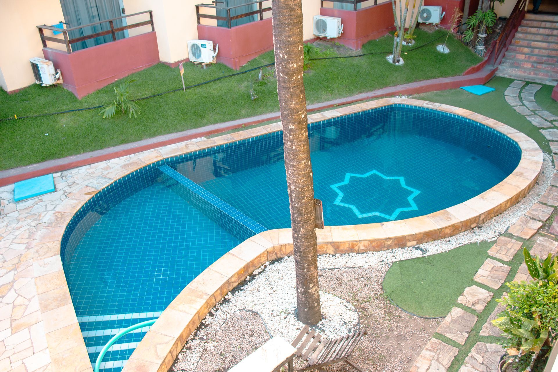 Piscina azul curva com uma palmeira no centro, rodeada por pavimentação em pedra e prédios de apartamentos.