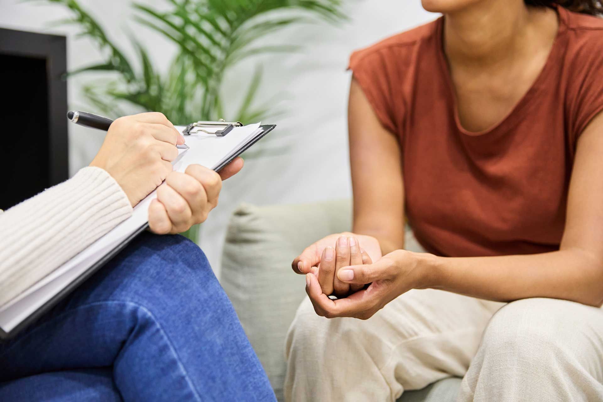A woman is sitting on a couch holding another woman 's hand while a person writes on a clipboard.