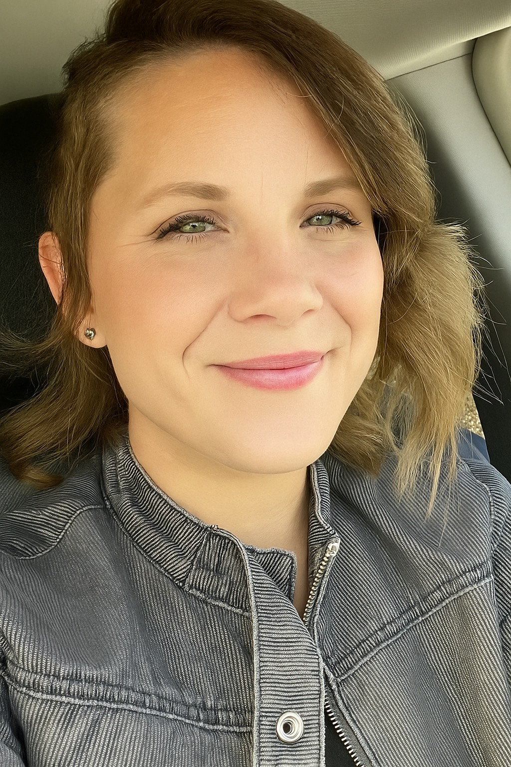 Woman with brown hair and denim jacket smiles in car.