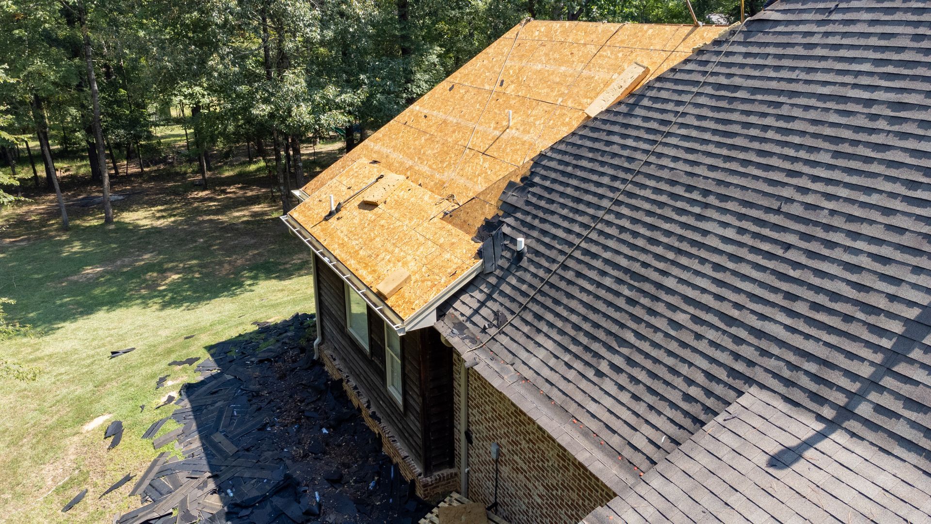 Roofing under repair on a house, with a new plywood section beside worn dark shingles.