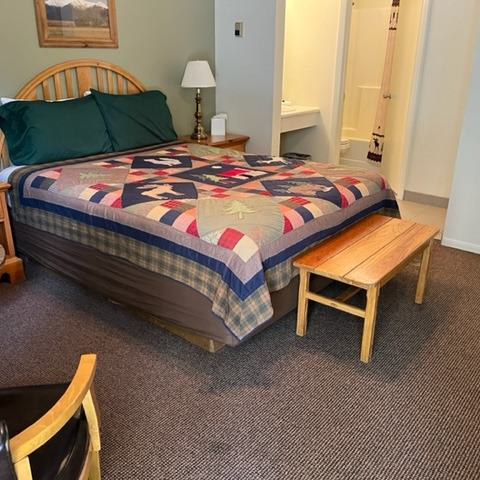 Hotel room with a quilt-covered bed, wooden bench, lamp, and bathroom in the background.