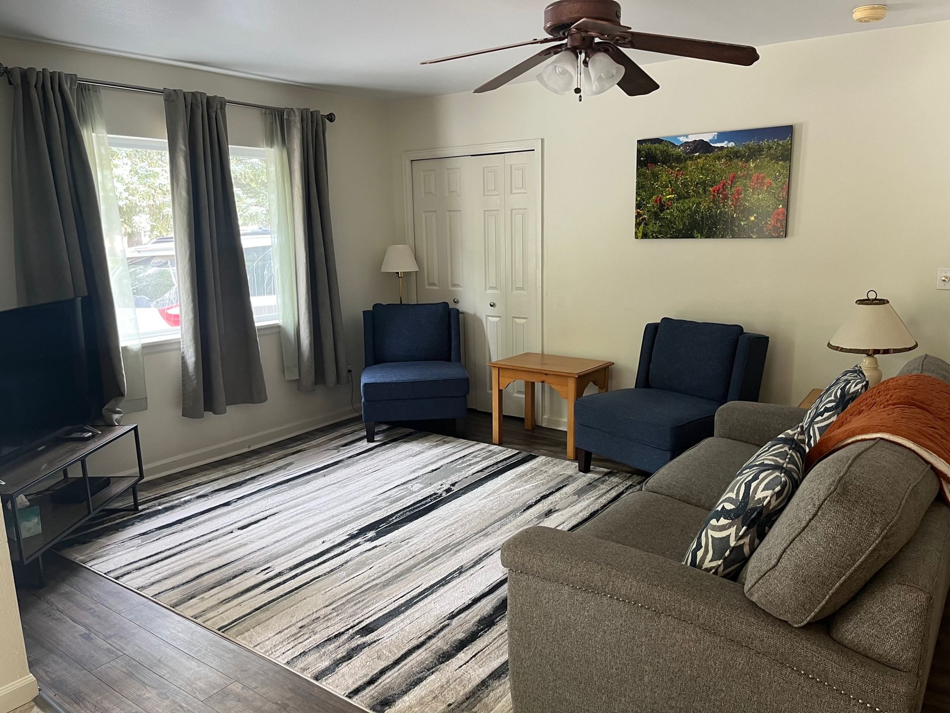 Living room with gray couch, blue chairs, rug, windows with curtains, and a ceiling fan.
