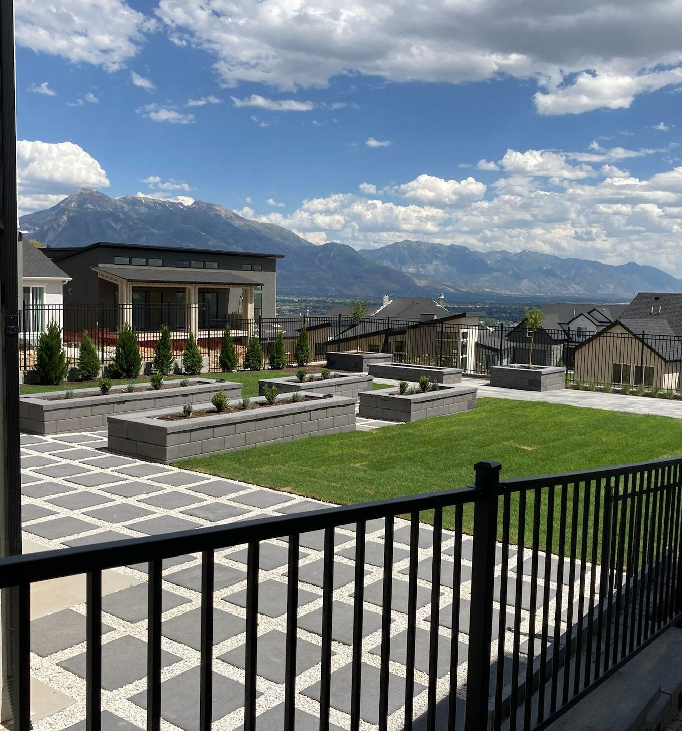 Patio with a grid of gray tiles and black railing overlooking a grassy area with raised garden beds and mountain view.