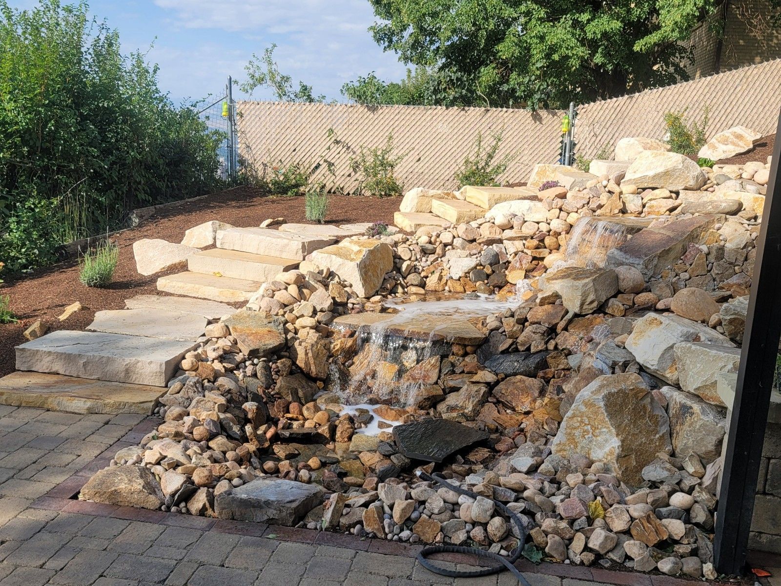 Waterfall feature with stone steps, built into a hillside. Water flows over rocks. Sunny outdoor setting.