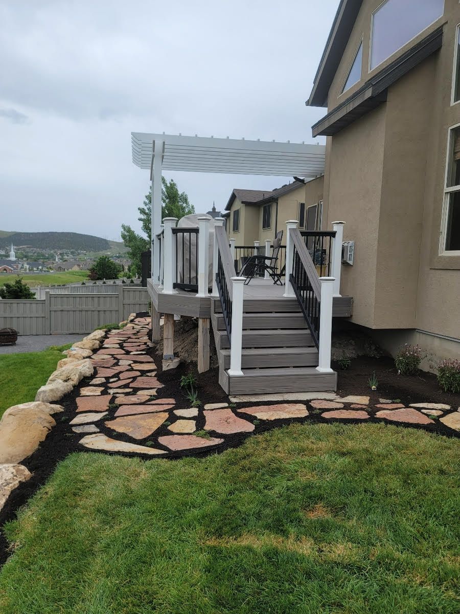 A deck with steps, a rock path, and a white pergola attached to a light brown house on a grassy lawn.