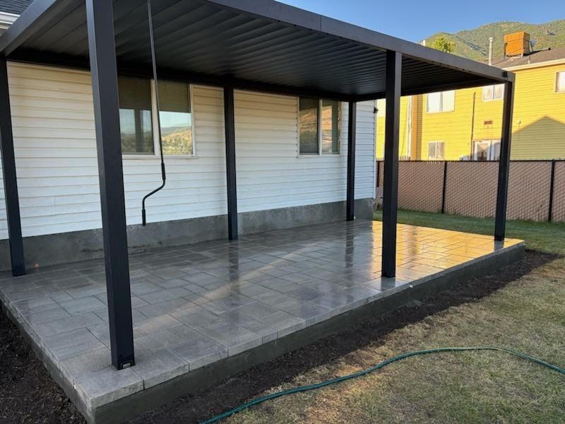 Black pergola covers a gray paved patio next to a white house, green grass on the ground.