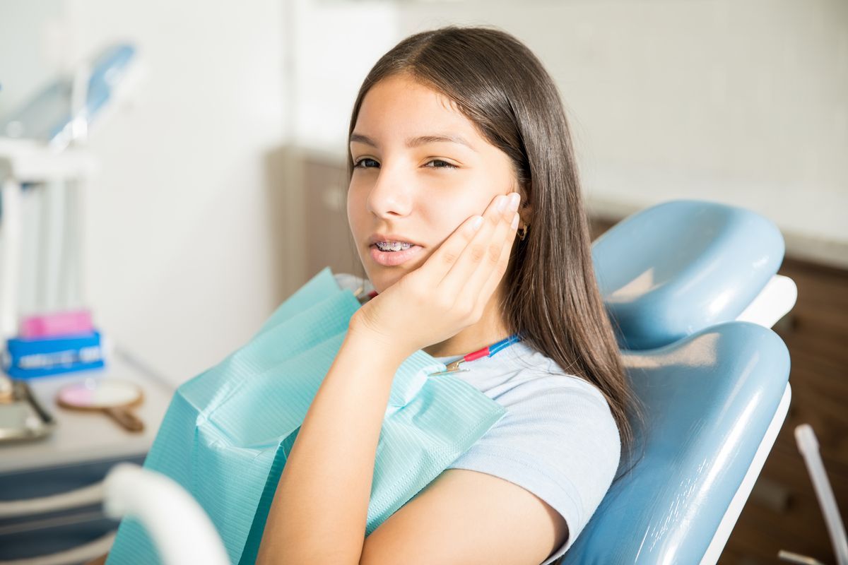 Young patient resting in a dental chair with a blue bib, touching her cheek in a clinic