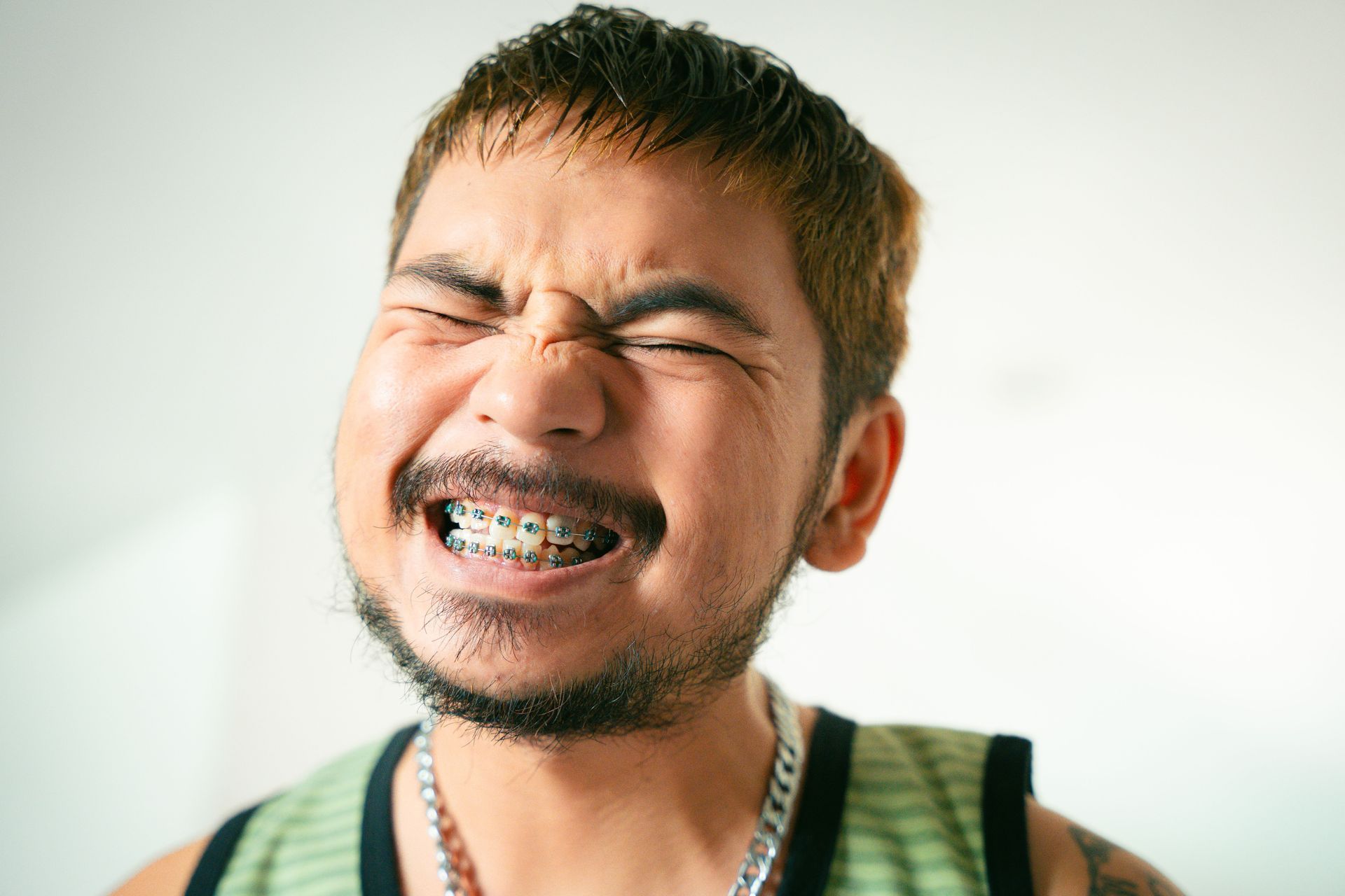 Guy with short hair in a green sleeveless shirt, showing braces and squinting his eyes